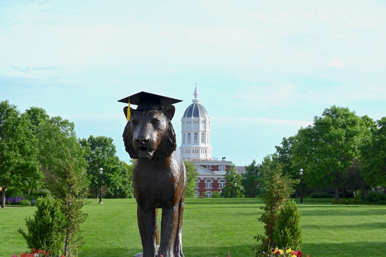 A photo of the bronze tiger on Canahan Quad with a graduation cap on top. Behind you can see Jesse Hall.
